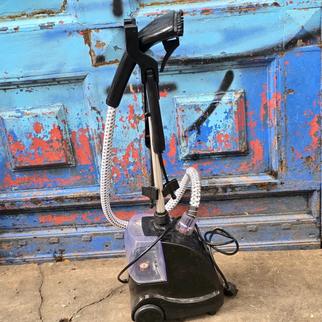 Vaporizador de ropa negro con tanque de agua transparente y cepillo, frente a una puerta azul desgastada. by Remates Narvaja