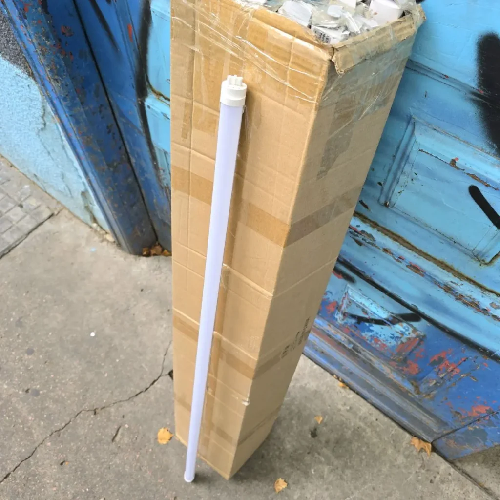 Tubo de luz LED blanco apoyado en una caja de cartón junto a una puerta azul desgastada. by Remates Narvaja