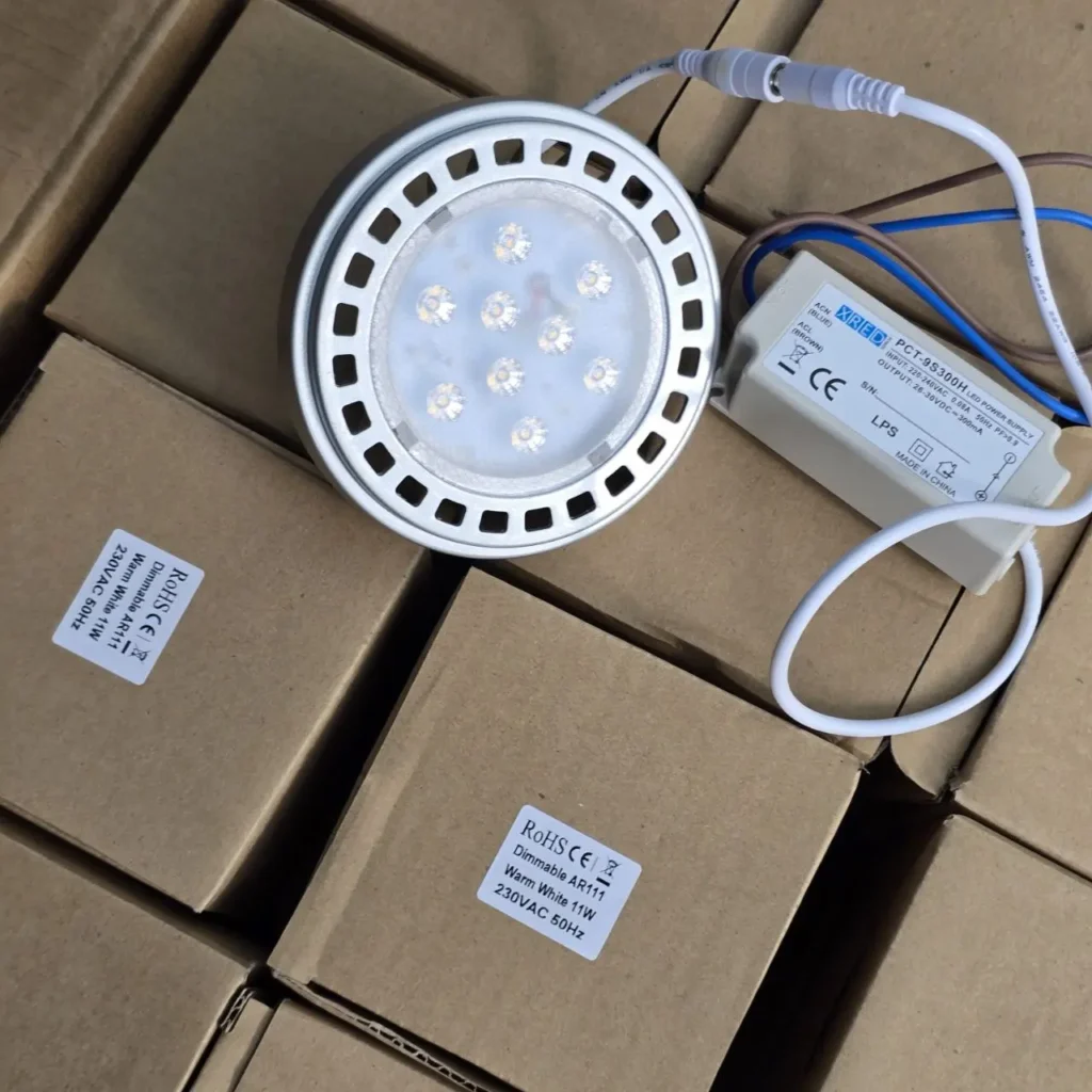 Lámpara LED AR111 regulable de 11W blanco cálido con fuente de alimentación sobre cajas de producto. by Remates Narvaja
