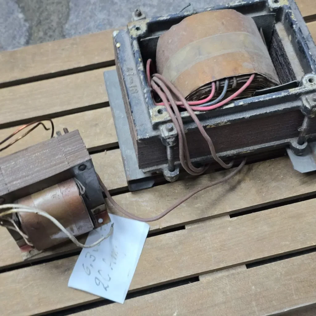Dos transformadores eléctricos vintage sobre una superficie de madera. El más grande tiene una bobina de cobre expuesta y cables, mientras que el más pequeño tiene una etiqueta escrita a mano que indica '6.3V 20 Amp'. by Remates Narvaja