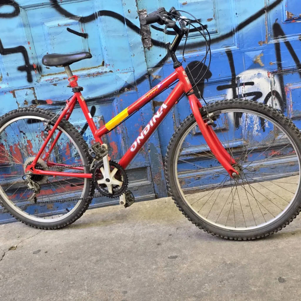Bicicleta de montaña roja ONDINA frente a puerta azul con graffiti." by Remates Narvaja