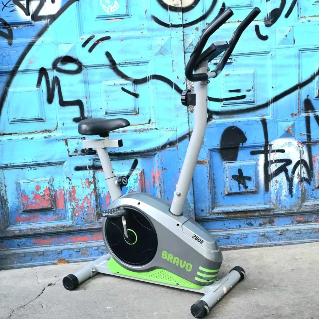 Bicicleta estática BRAVO 280E gris y verde lima frente a una puerta azul con graffiti. by Remates Narvaja