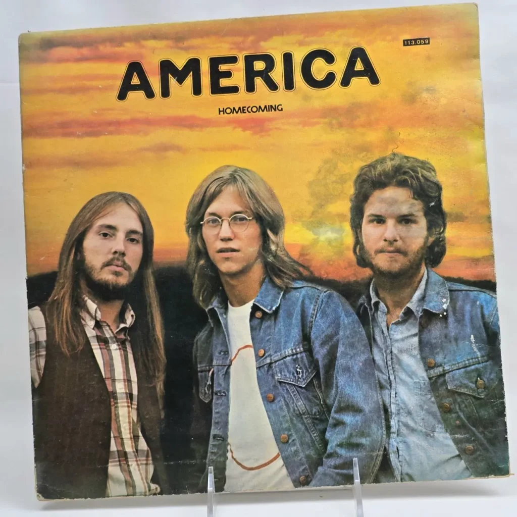Portada del álbum "Homecoming" de America, con los tres miembros de la banda de pelo largo, vestidos con mezclilla y cuadros, frente a un by Remates Narvaja