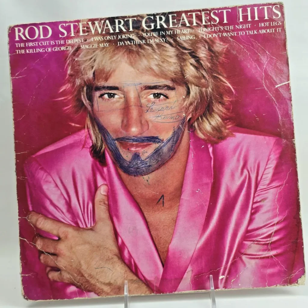 Portada del álbum Rod Stewart Greatest Hits con Rod Stewart en chaqueta rosa, garabatos de barba y gafas en su cara, y desgaste visible. by Remates Narvaja