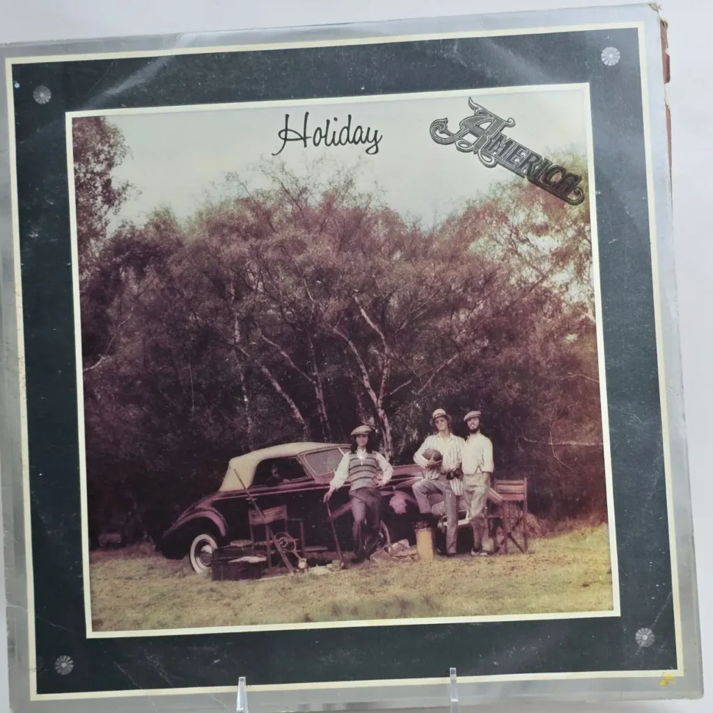 Portada del álbum "Holiday" de America, con tres hombres posando junto a un coche descapotable vintage en un entorno boscoso by Remates Narvaja
