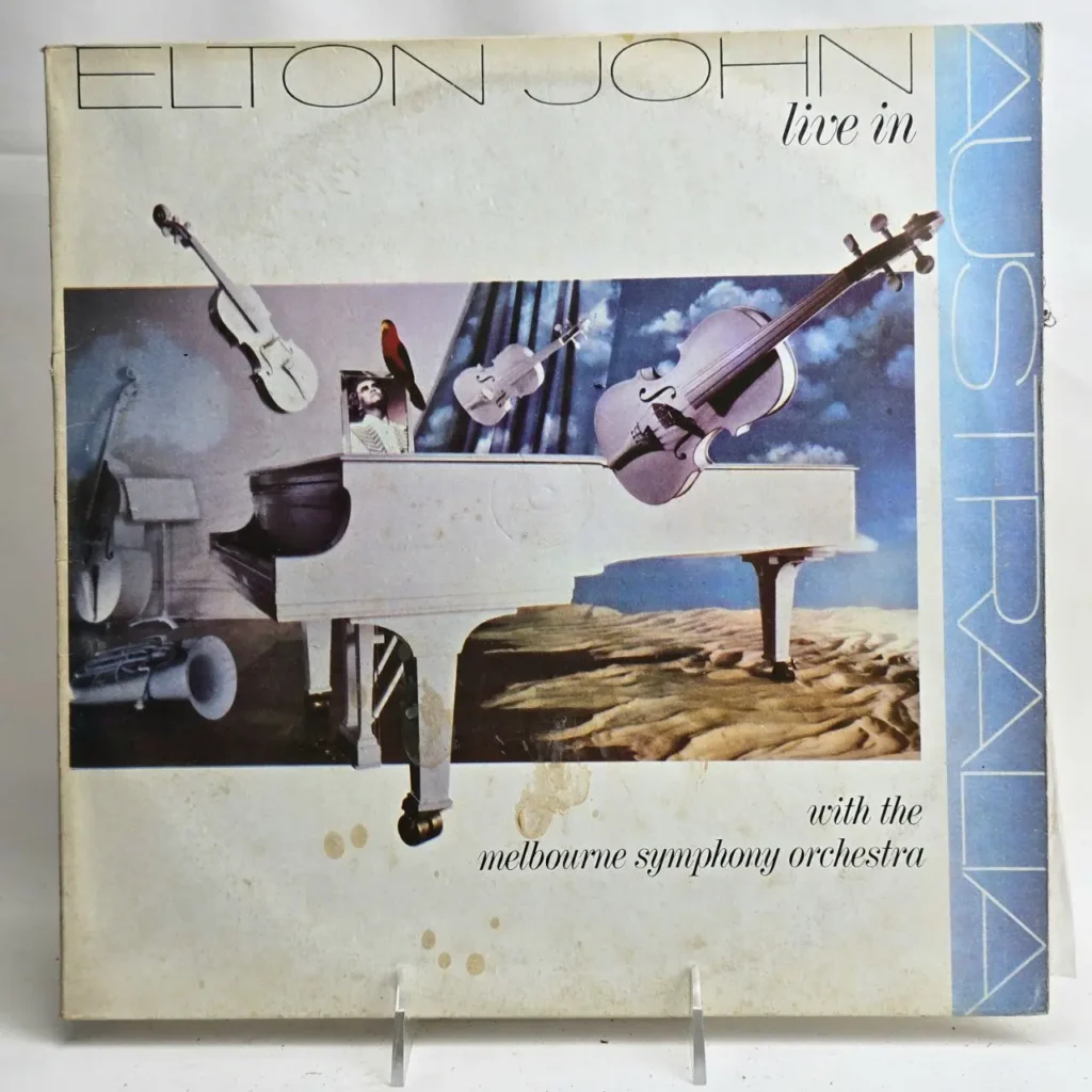 Portada del álbum Elton John Live in Australia con la Melbourne Symphony Orchestra, mostrando un piano blanco, violines flotantes y un paisaje desértico, estilo by Remates Narvaja