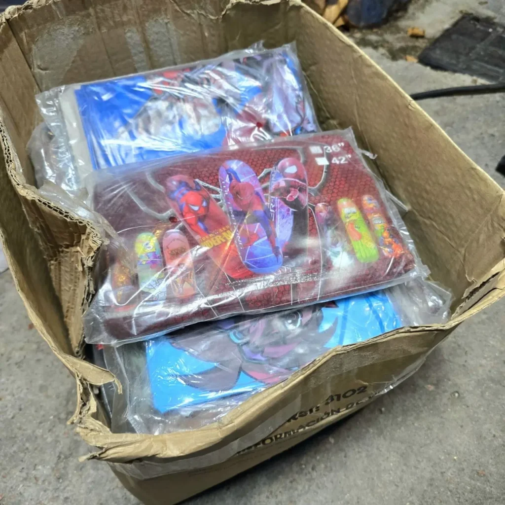 Caja de cartón llena de productos envueltos individualmente con temática de Spider-Man y otros personajes de dibujos animados, posiblemente ropa de cama o artículos by Remates Narvaja