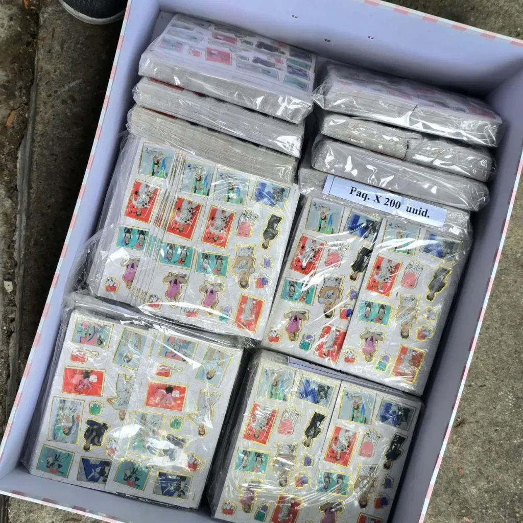 Caja abierta llena de paquetes individuales de hojas de pegatinas decorativas con diseños de dibujos animados. Una etiqueta indica 'Paq. X 200 unid.'. by Remates Narvaja