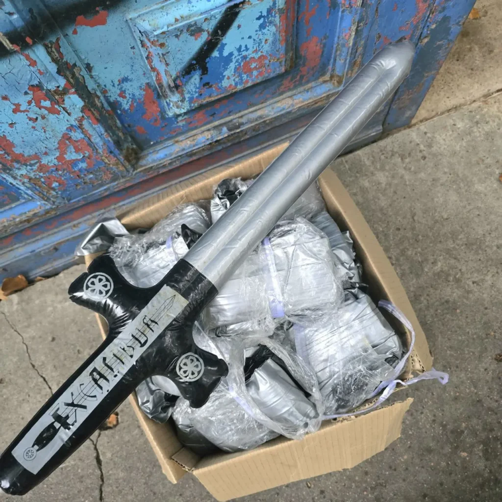 Espada inflable Excalibur plateada y negra sobre una caja llena de más espadas de juguete. by Remates Narvaja