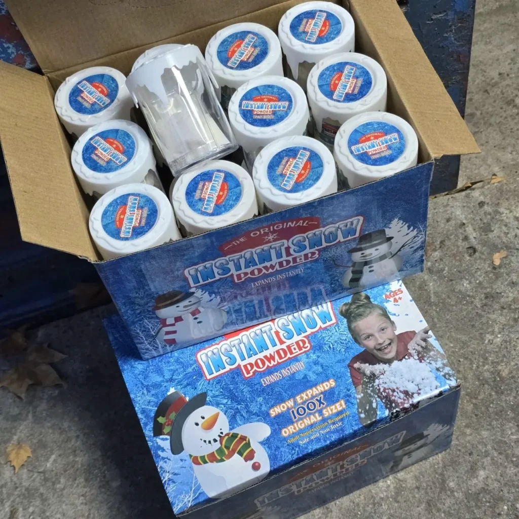 Caja abierta llena de múltiples recipientes pequeños de "The Original Instant Snow Powder" con etiquetas azules, apilada sobre otra caja de exhibición de la by Remates Narvaja