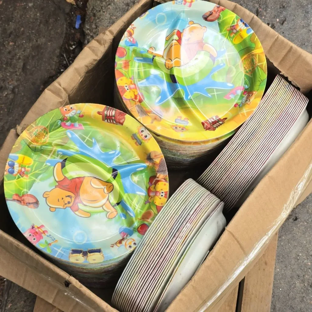 Platos desechables coloridos con personajes de Winnie the Pooh para fiestas infantiles, apilados en una caja de cartón junto a platos blancos by Remates Narvaja