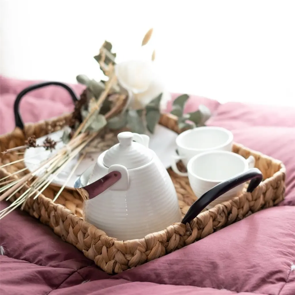 Bandeja de mimbre con tetera blanca, tazas y flores secas sobre una cama rosa. by Remates Narvaja