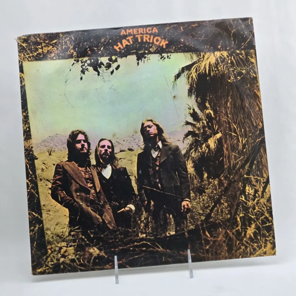 Portada del álbum de vinilo "America Hat Trick" con los tres miembros de la banda en un paisaje desértico con palmeras. by Remates Narvaja