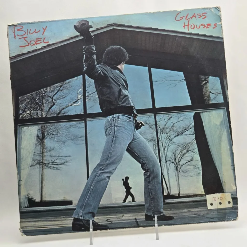 Portada del álbum Glass Houses de Billy Joel, con el cantante en chaqueta de cuero a punto de lanzar una piedra a un edificio de cristal. by Remates Narvaja