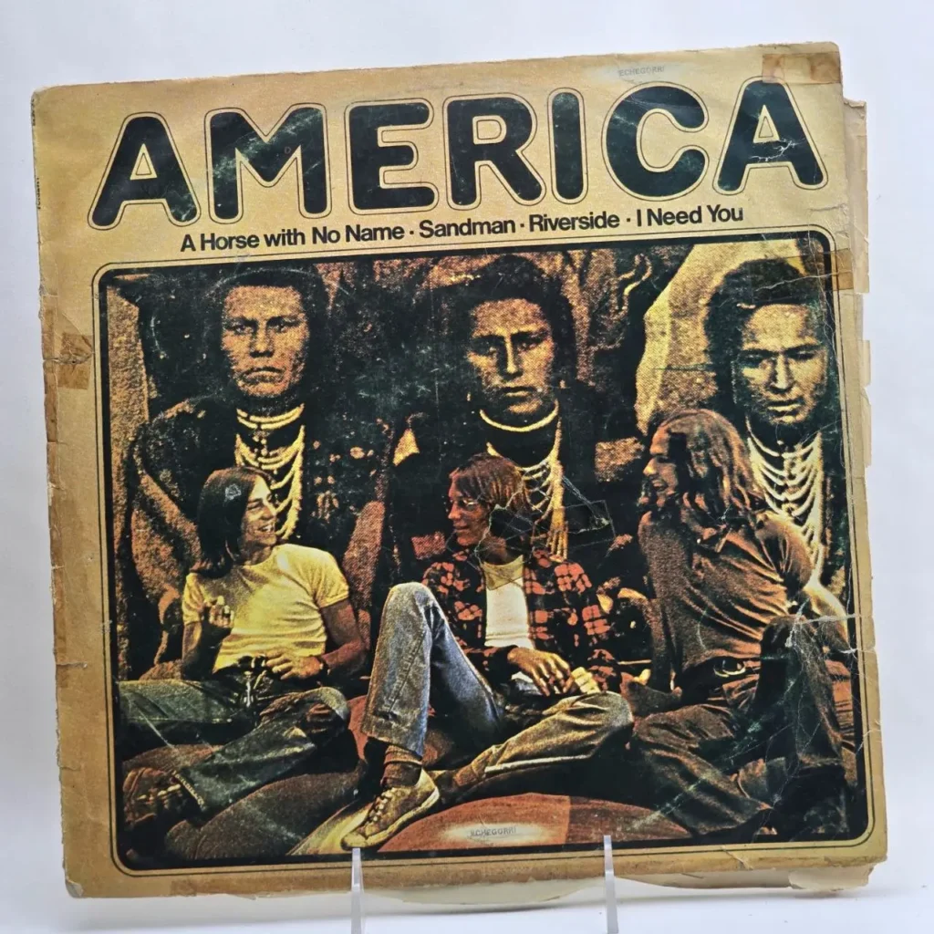Portada del álbum "America" con el título y canciones como "A Horse with No Name", mostrando a los tres miembros de la banda sentados frente a figuras by Remates Narvaja