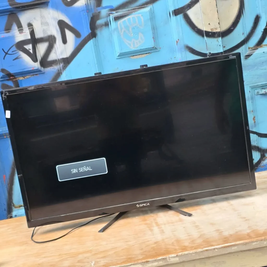 Televisor de pantalla plana negro marca Spica mostrando el mensaje "SIN SEÑAL" sobre una mesa de madera, con una pared azul grafitada de fondo. by Remates Narvaja