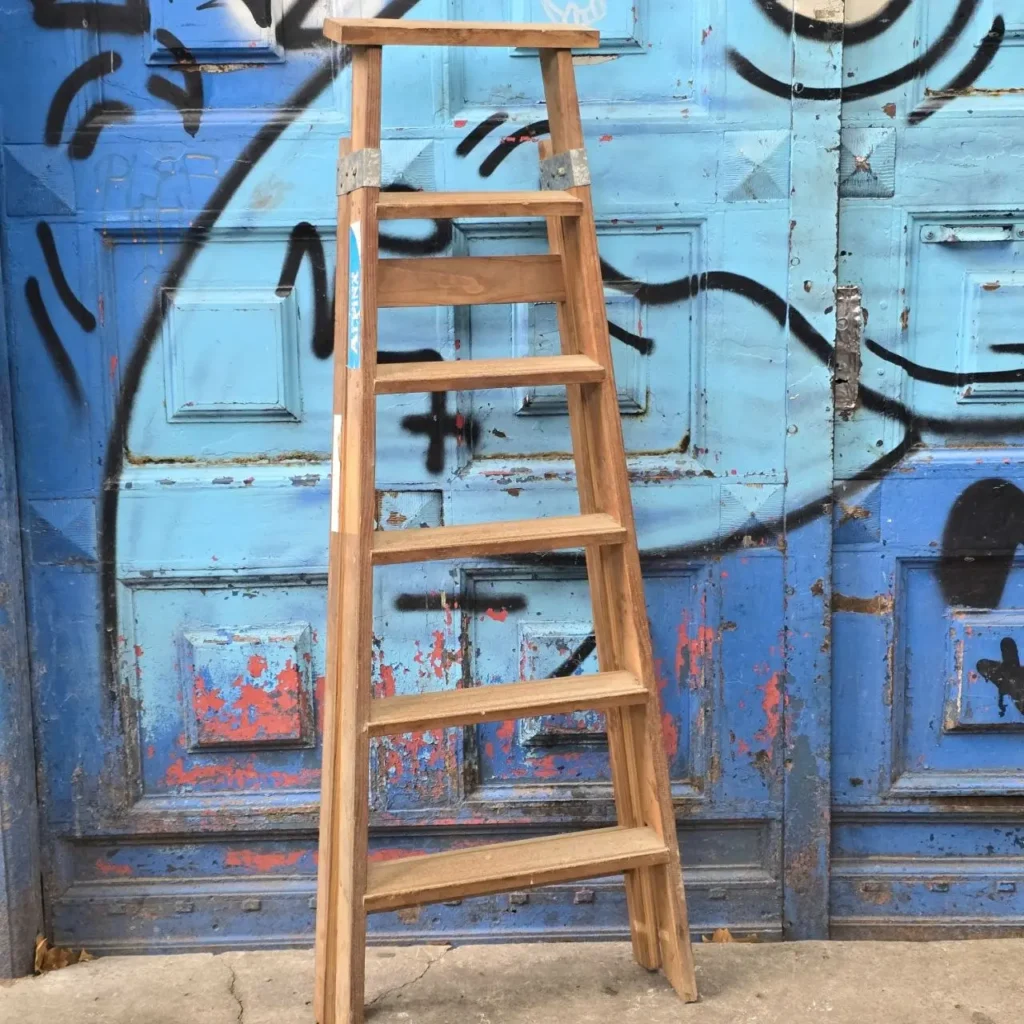 Escalera de madera apoyada en una puerta azul desgastada con graffiti. by Remates Narvaja