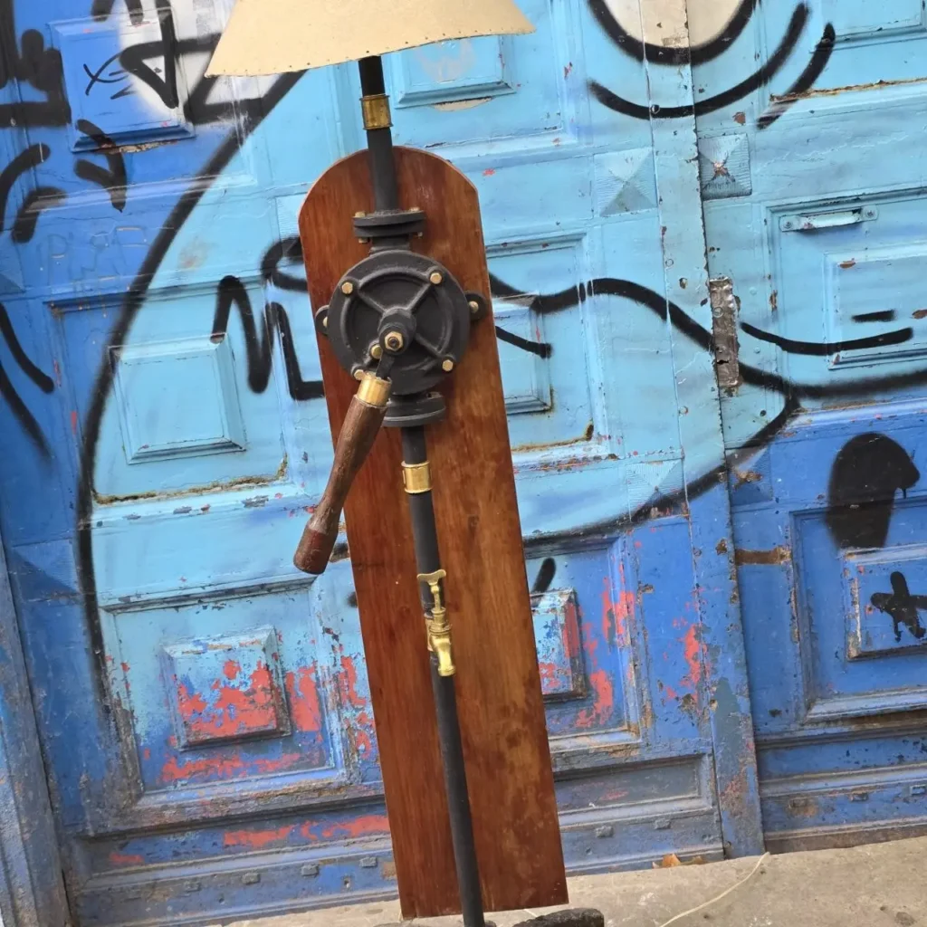 Lámpara de pie industrial con mecanismo de bomba, madera y metal, frente a puerta azul con graffiti. by Remates Narvaja