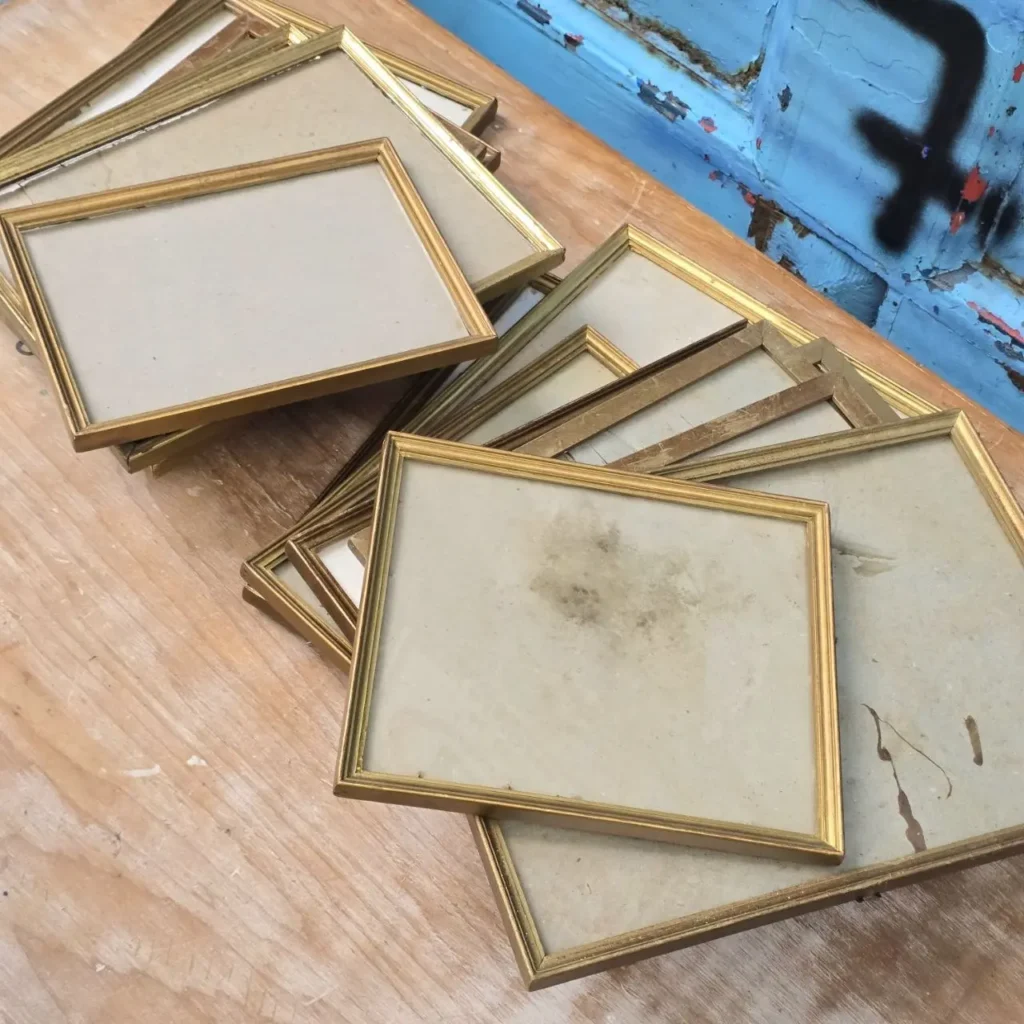 Marcos de fotos dorados vintage vacíos apilados sobre una superficie de madera clara, con una pared azul desgastada al fondo. by Remates Narvaja