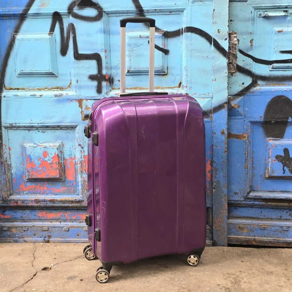 Maleta morada con ruedas frente a una puerta azul desgastada con graffiti. by Remates Narvaja