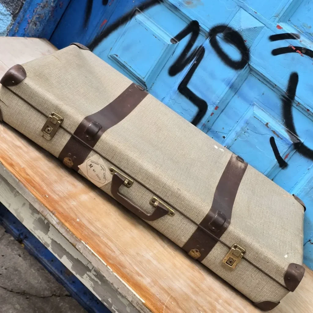 Estuche vintage beige con correas de cuero marrón sobre madera y fondo azul con grafitis. by Remates Narvaja