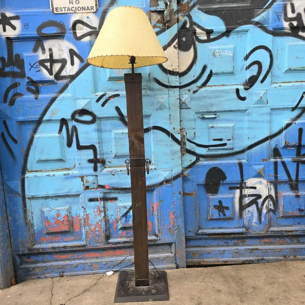 Lámpara de pie con pantalla beige y poste oscuro frente a una puerta azul graffiteada. by Remates Narvaja