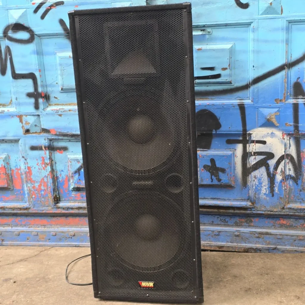 Altavoz Novik negro con doble woofer y rejilla metálica, frente a una puerta azul con grafitis. by Remates Narvaja