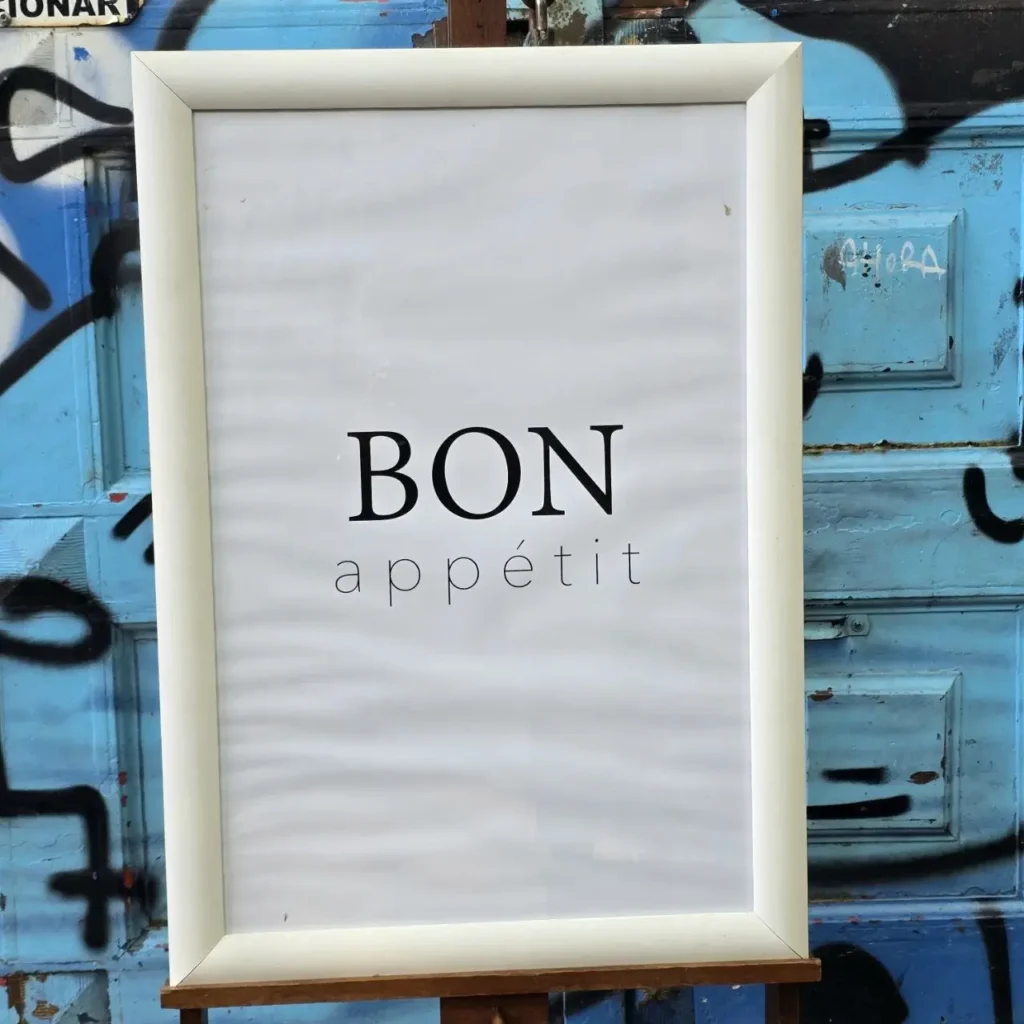 Cartel blanco con la frase "BON appétit" sobre un fondo azul con grafitis. by Remates Narvaja