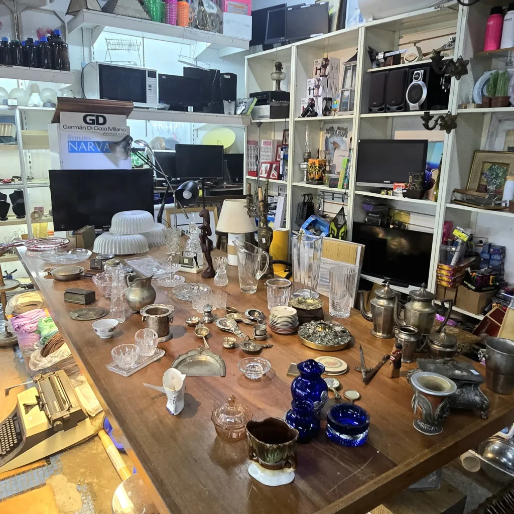 Mesa de remates con antigüedades, cristalería, objetos de metal y electrónica en estanterías. by Remates Narvaja