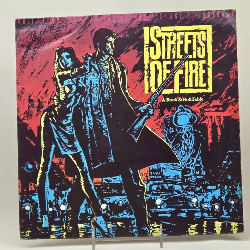 Streets of Fire soundtrack album cover con arte estilizado de un hombre con escopeta y una mujer, con fondo rojo y paisaje urbano azul. by Remates Narvaja