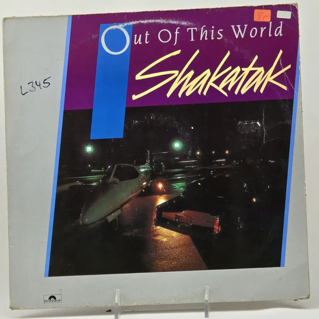 Portada del álbum "Out Of This World" de Shakatak, con un jet privado y un coche de lujo en una escena nocturna. by Remates Narvaja