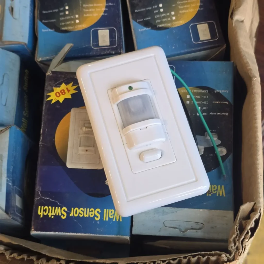 Un interruptor de sensor de movimiento de pared blanco con un indicador LED verde y un botón, sobre cajas de producto azules en una caja de cartón. by Remates Narvaja