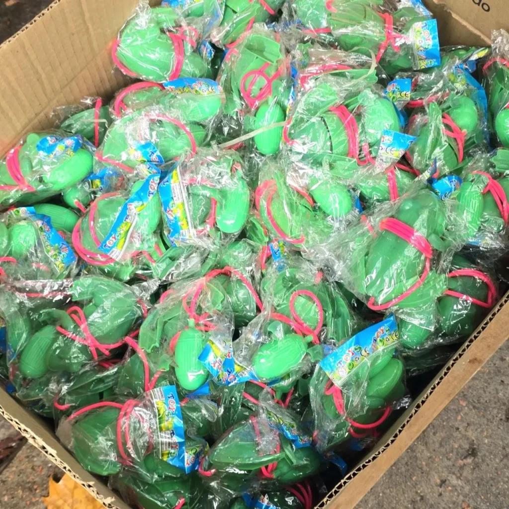 Caja llena de pequeños juguetes de plástico verdes envueltos individualmente con bandas rosas. by Remates Narvaja