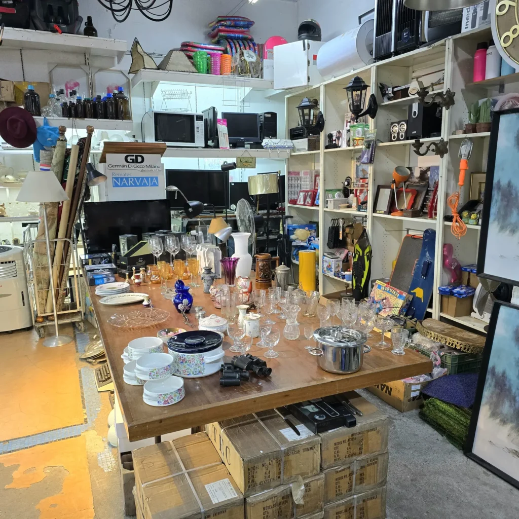 Local de remates o tienda de segunda mano con una mesa central llena de cristalería y vajilla, rodeada de estanterías con electrónica, artículos by Remates Narvaja