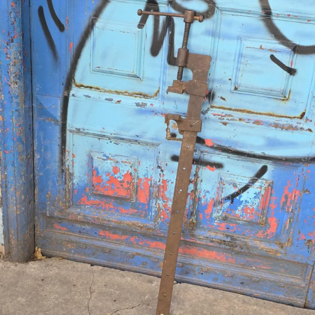 Sargento de carpintero oxidado apoyado en una puerta azul antigua con pintura descascarada y grafitis. by Remates Narvaja