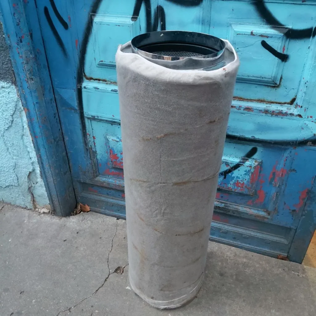 Filtro cilíndrico aislado con tapa metálica frente a puerta azul de madera con graffiti. by Remates Narvaja