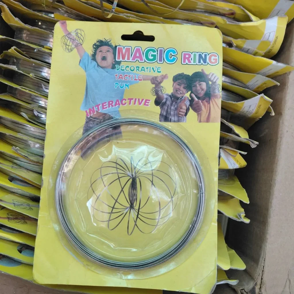 Juguete cinético "Magic Ring" de metal en espiral en su empaque amarillo, con niños jugando y otros paquetes apilados by Remates Narvaja