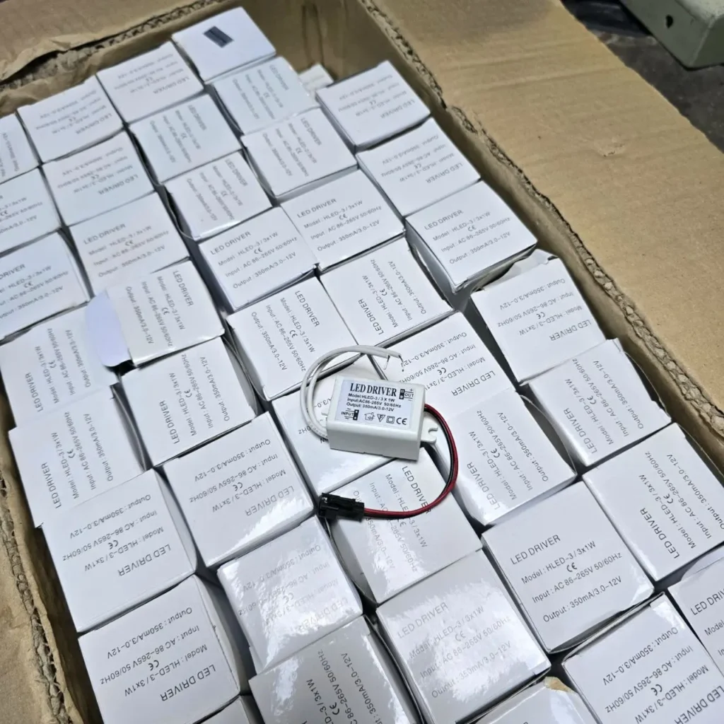 Caja de cartón llena de controladores LED (LED drivers) en sus cajas blancas, con un controlador HLED-3/3x1W suelto visible. by Remates Narvaja