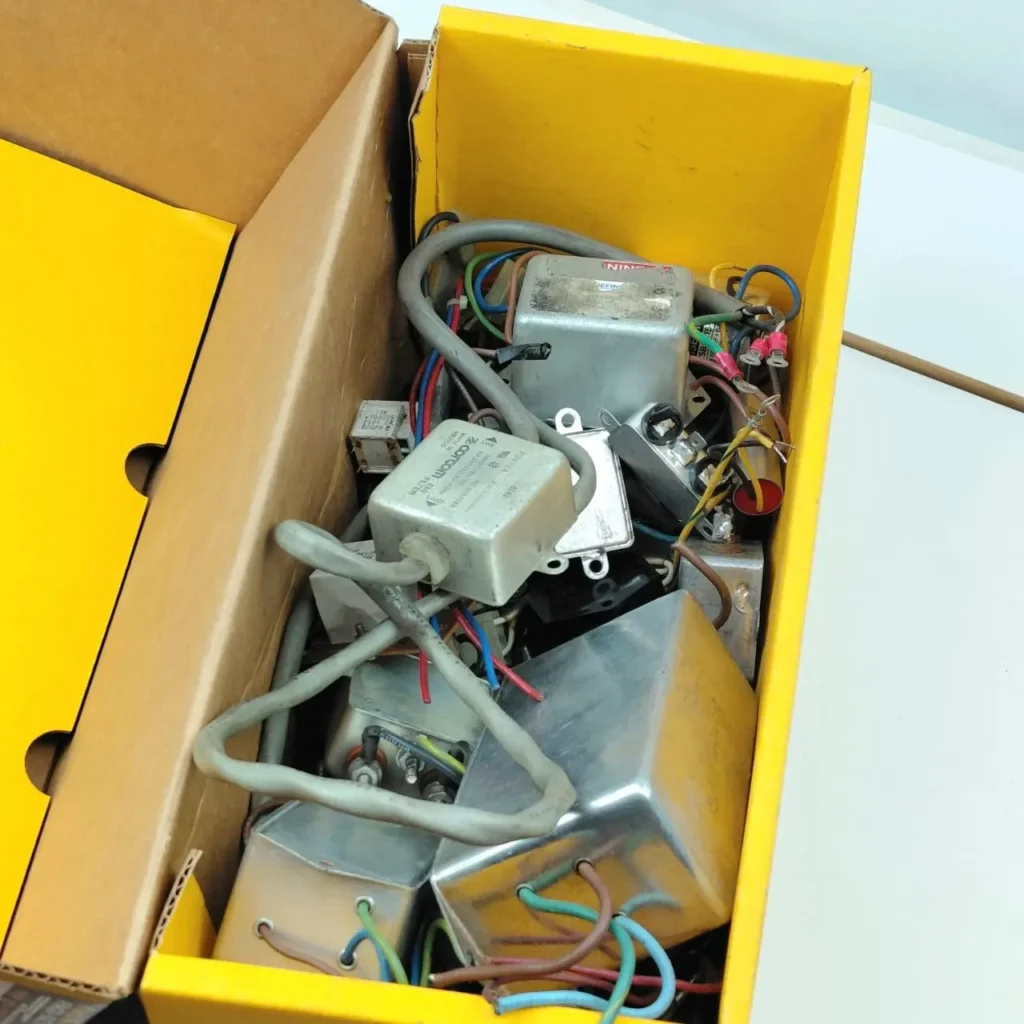 Caja amarilla llena de componentes electrónicos variados, incluyendo transformadores, filtros y cables. by Remates Narvaja