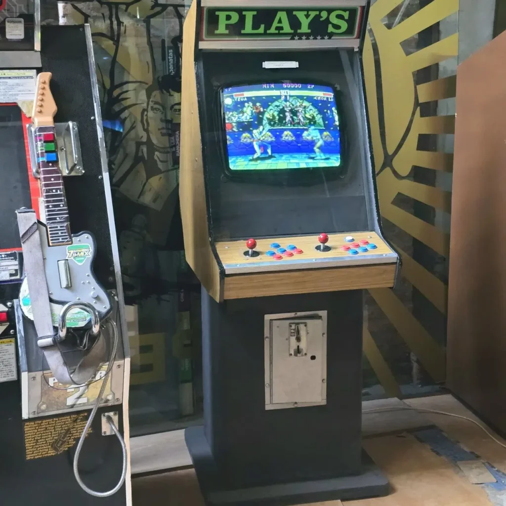 Máquina arcade retro "PLAY'S" con un juego de lucha en pantalla y joysticks rojos, junto a una máquina de juego de guitarra Z by Remates Narvaja
