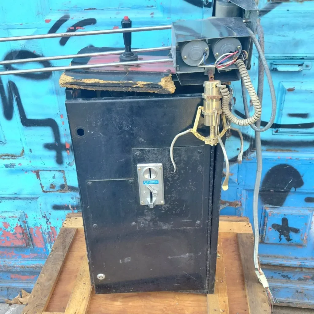Máquina de garra (claw machine) vintage o averiada con ranura para monedas y joystick, sobre palets de madera. by Remates Narvaja