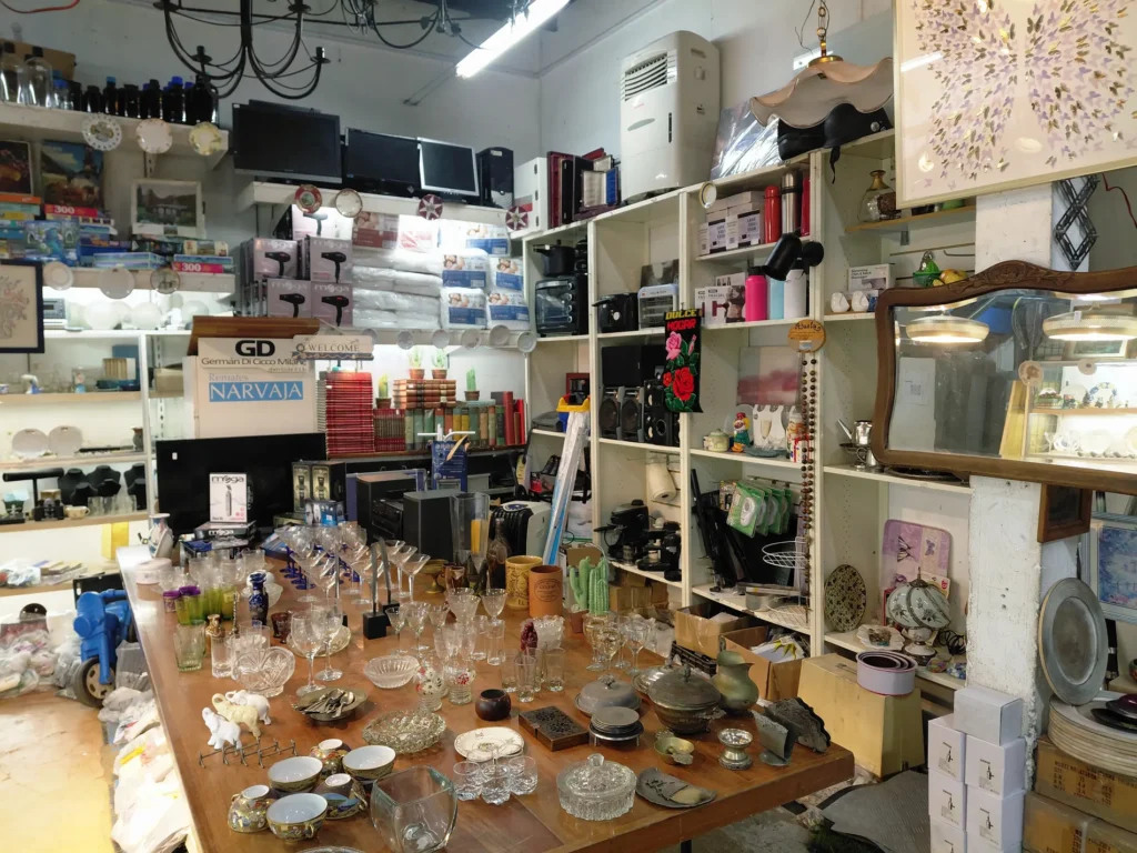 Tienda de segunda mano o remates con estanterías llenas de electrónica, libros y artículos para el hogar, y una mesa grande con cristaler by Remates Narvaja