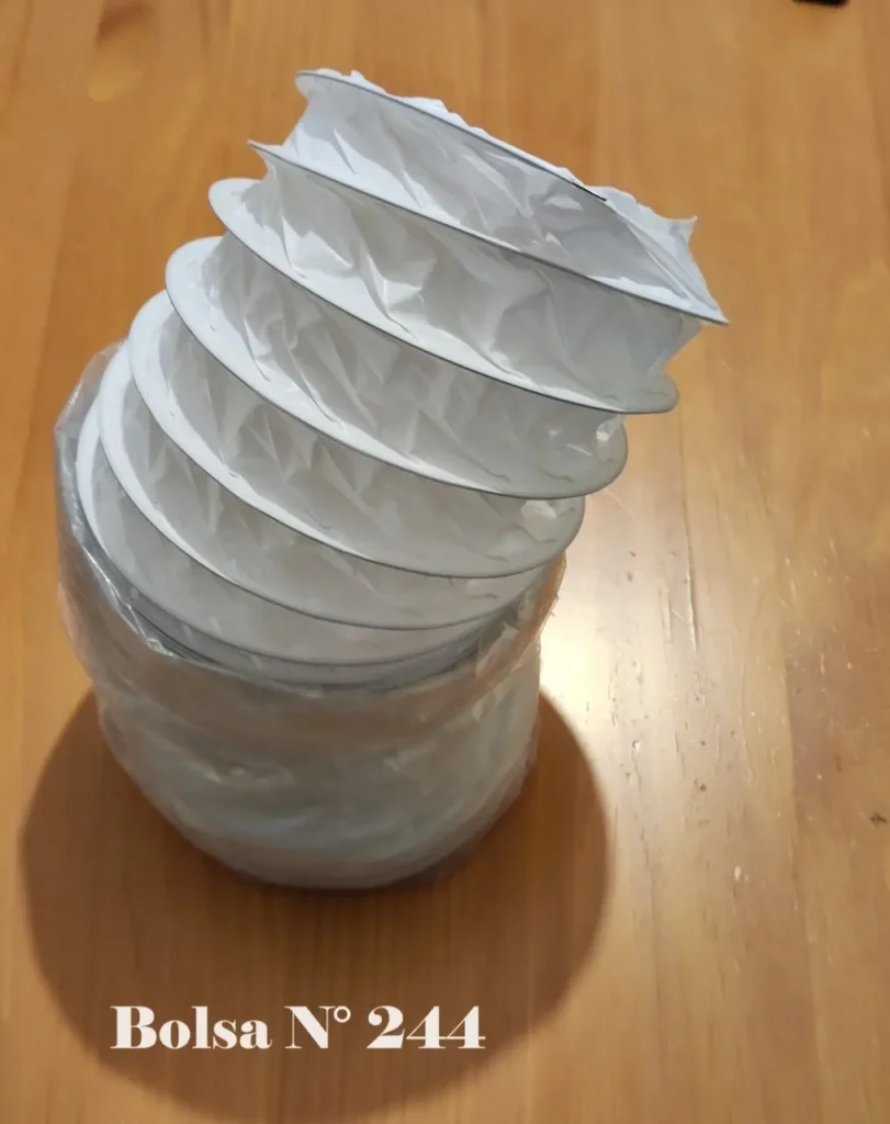 Tubo flexible de ventilación blanco tipo acordeón, parcialmente comprimido, en una bolsa plástica sobre madera. Incluye el texto "Bols by Remates Narvaja