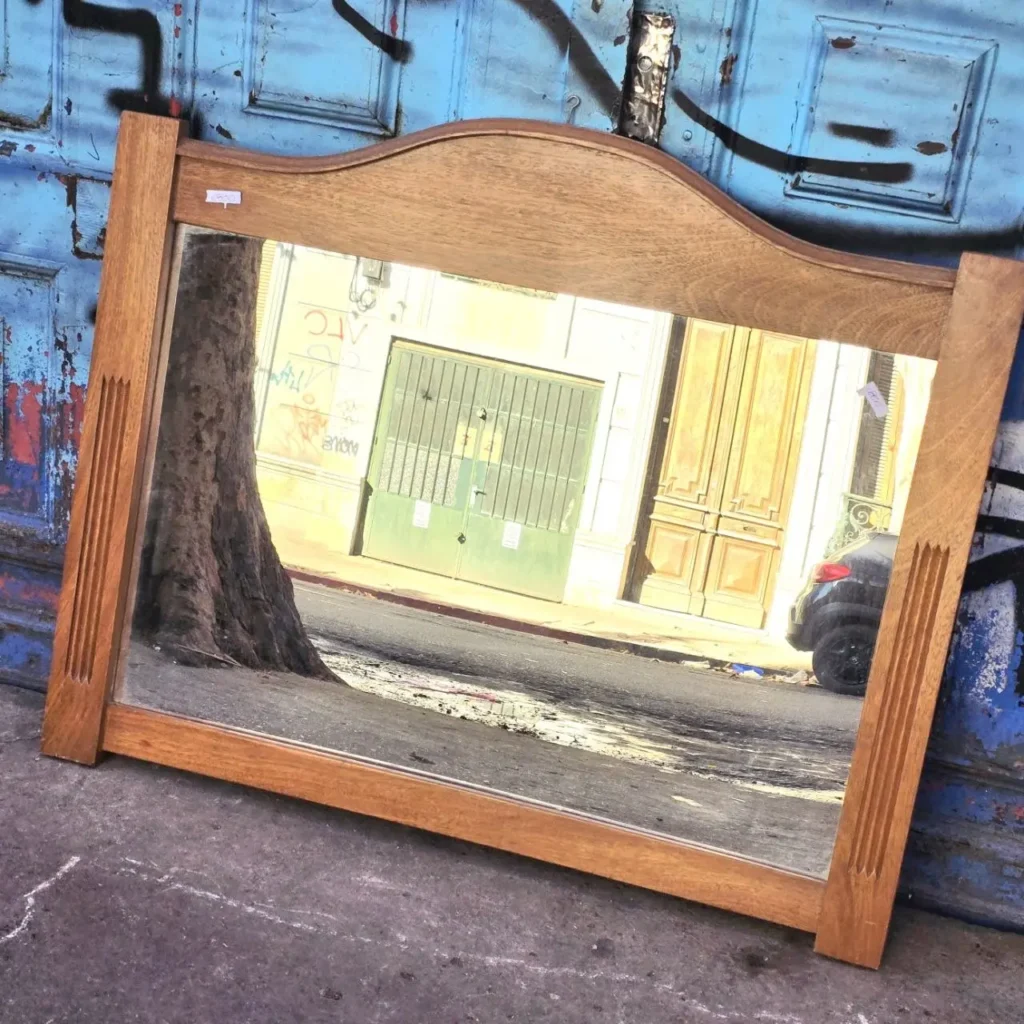 Espejo de madera vintage con marco decorado, apoyado en una pared azul y reflejando una calle urbana. by Remates Narvaja