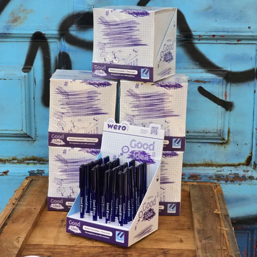 Expositor de bolígrafos borrables Good Idea de tinta azul, con cajas de producto y fondo rústico azul. by Remates Narvaja