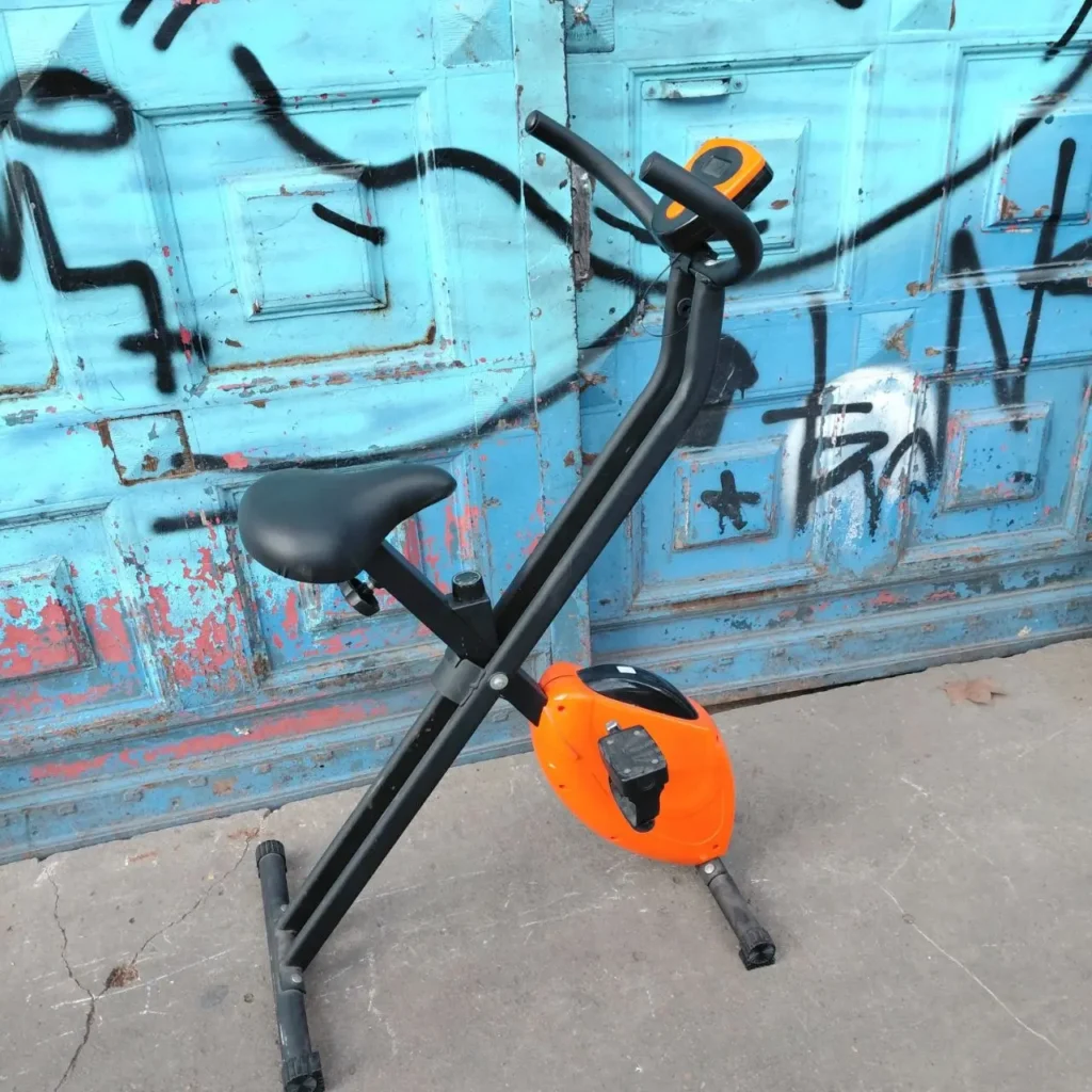 Bicicleta estática negra y naranja frente a una puerta azul grafiteada. by Remates Narvaja
