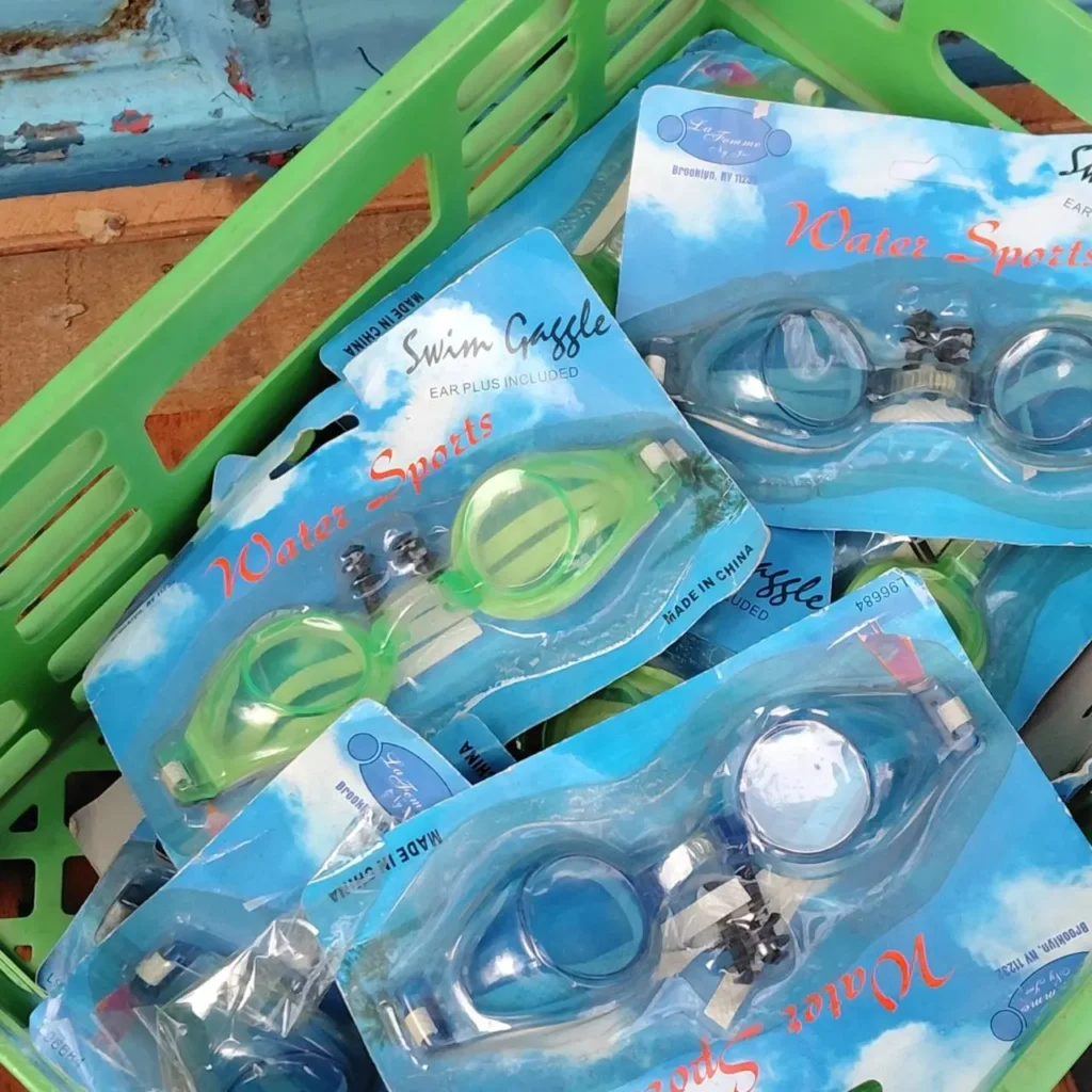 Múltiples paquetes de gafas de natación azules y verdes, etiquetadas como 'Water Sports Swim Gaggle' con tapones para los oídos incluidos, apiladas en una cesta de plástico verde. by Remates Narvaja