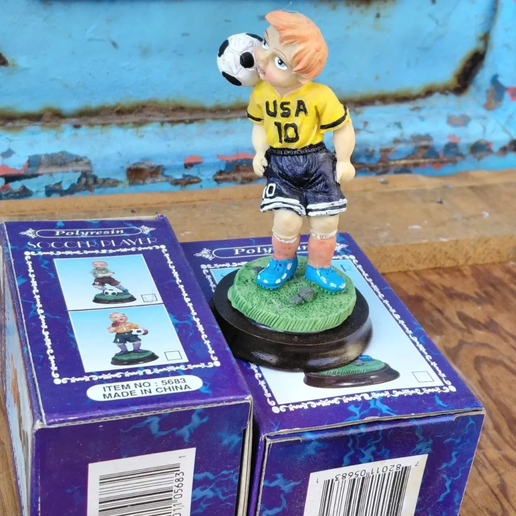 Figurilla de jugador de fútbol de poliresina con camiseta amarilla de USA #10, equilibrando un balón en la cabeza, sobre dos cajas moradas. by Remates Narvaja