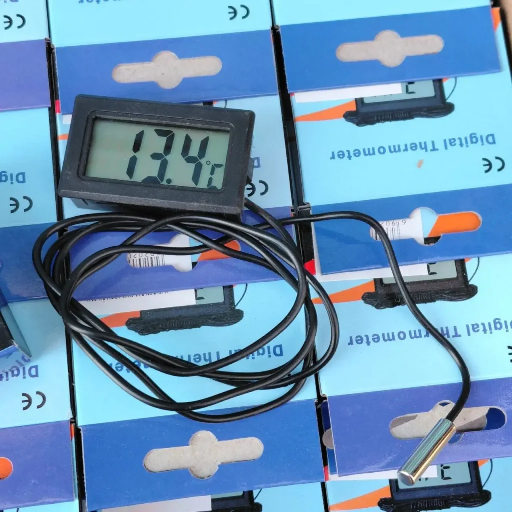 Termómetro digital negro con sonda metálica mostrando 13.4°C sobre cajas de embalaje azules. by Remates Narvaja