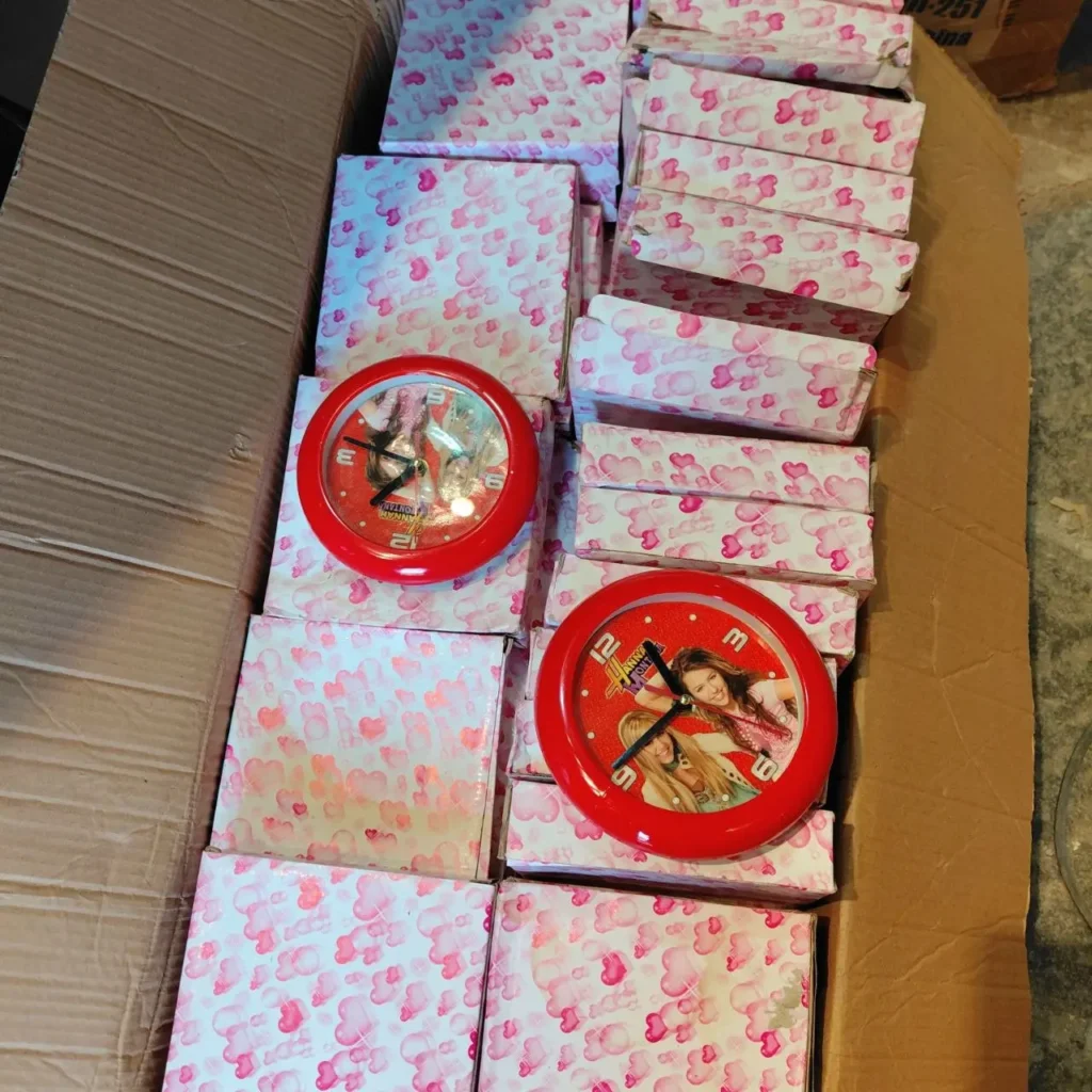 Caja de cartón llena de pequeñas cajas envueltas en papel con patrón de corazones rosas y dos relojes redondos rojos con imágenes de mujeres jóvenes en sus esferas. by Remates Narvaja